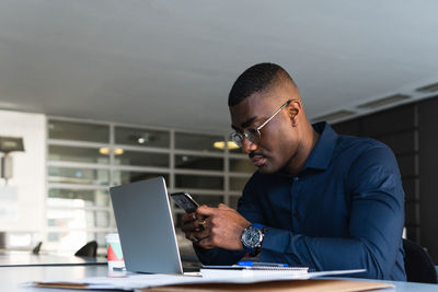 Side view of african american concentrated male in eyeglasses and formal clothes sitting in chair at table while using phone and netbook near document and takeaway drink in light workspace