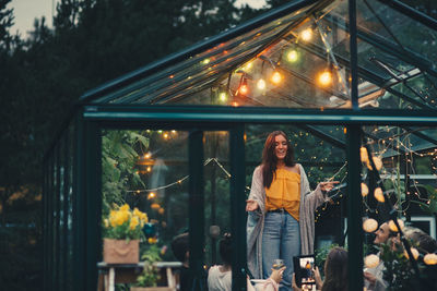 Smiling young woman standing amidst friends in glass conservatory at back yard