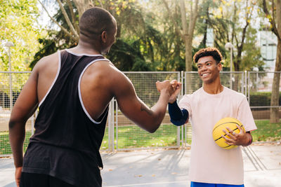 Young ethnic back male athletes with yellow basketball shaking hands while standing on sports ground in park looking at each other