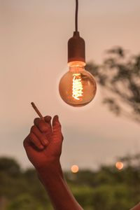 Cropped image of hand holding cigarette against illuminated light bulb during sunset