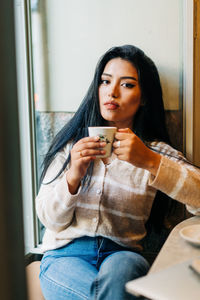 Ethnic female with makeup drinking hot beverage from cup in coffee house