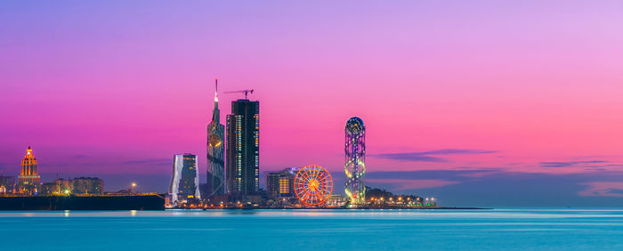 Illuminated city by sea against sky at sunset