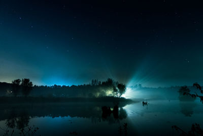 Reflection of silhouette trees on calm lake against star field
