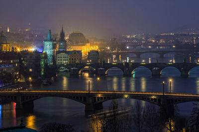 Illuminated bridge over river by buildings against sky at night