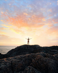 Distant view of person standing on rocky shore against cloudy sky during sunset