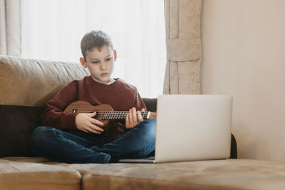 Boy playing guitar while sitting on sofa at home