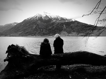 Rear view of mother and daughter leaning on log by lake against sky