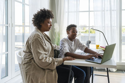 Mother sitting by son doing homework using laptop on table at home