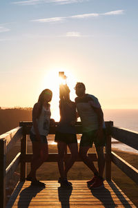 Smiling tourists leaning on wooden railing and taking self portrait against sunset sky over calm sea