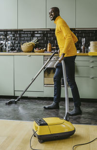 Full length of smiling man using vacuum cleaner doing chores in kitchen at home