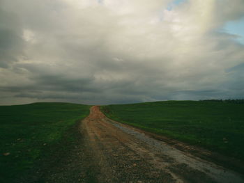 Road passing through grassy field against cloudy sky
