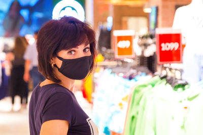 A woman in a black protective mask in a shopping mall among hangers with things during the pandemic.