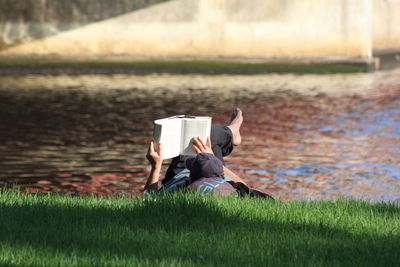 Man reading book while relaxing on grassy field by lake