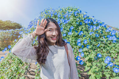 Portrait of young woman raising hand up while standing against flowering plants during sunny day