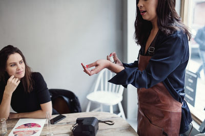 Midsection of woman spraying perfume on wrist by female colleague sitting at table in workshop