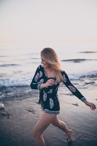 Happy young woman running at beach against sky during sunset