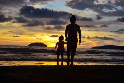 Rear view of silhouette man with daughter on shore at beach during sunset