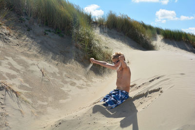 Full length of shirtless boy on beach. sitting in the dunes and play with sand