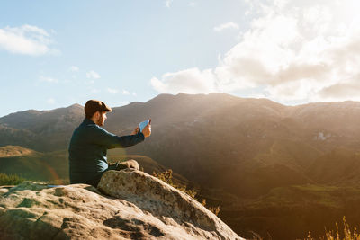 Man sitting on rock looking at mountains against sky