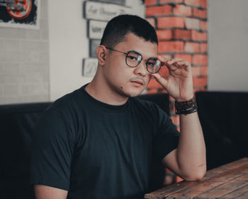 Portrait of young man holding eyeglasses against wall