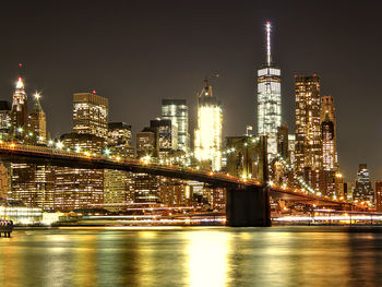 Illuminated modern buildings by river against sky at night