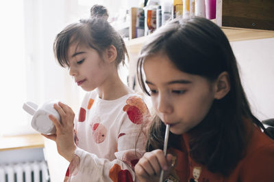 Girl drinking by sister looking at mug in kitchen