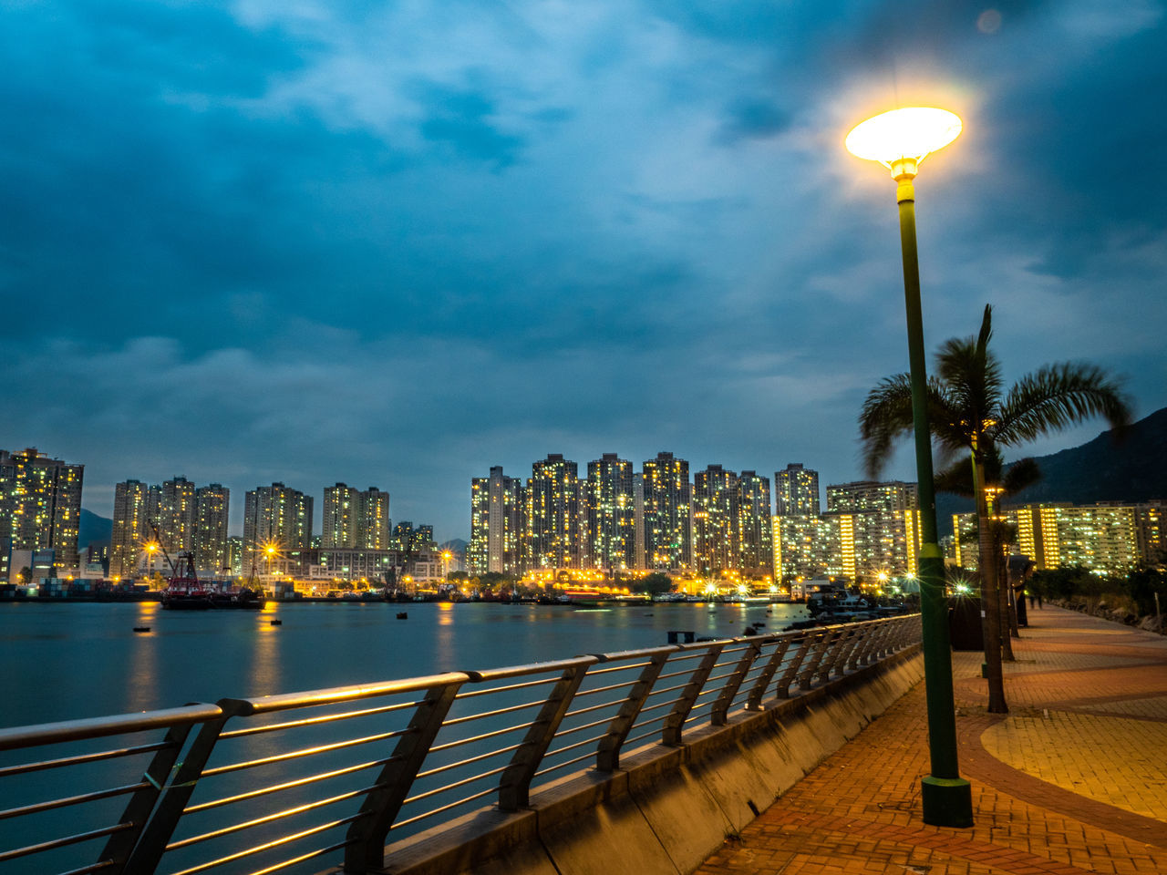 illuminated, architecture, building exterior, sky, city, built structure, water, cloud - sky, palm tree, tropical climate, lighting equipment, building, cityscape, railing, night, street light, tree, nature, dusk, office building exterior, skyscraper, outdoors, no people, light