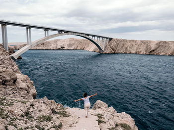 Woman standing on sea shore, looking at arched bridge across sea channel.