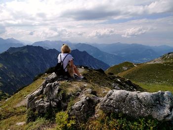Rear view of woman sitting on rock looking at mountains against sky