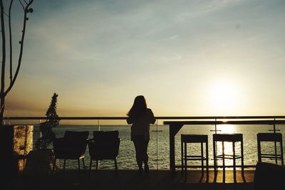 Rear view of woman standing by railing against sunset sky