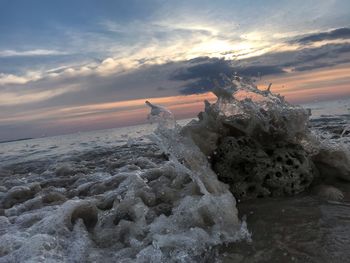 Sea waves splashing on rocks against sky during sunset