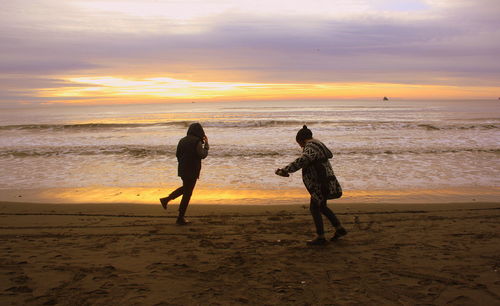 Friends running at beach against cloudy sky during sunset