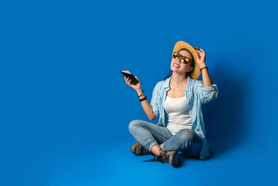 Young woman using smart phone while sitting on blue wall