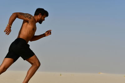 Shirtless young man running on sand at desert during sunny day