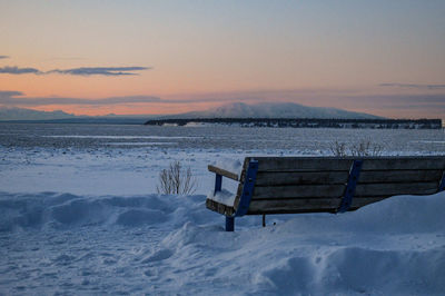 Empty bench on snow covered shore against sky during sunset
