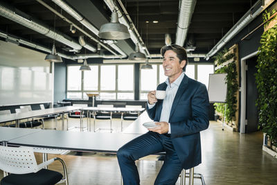 Smiling businessman with cup of coffee sitting on table in meeting room of an office