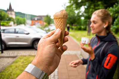 Cropped hand of man holding ice cream cone against woman standing on footpath in background