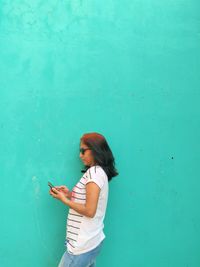 Side view of young woman using phone while standing against blue wall