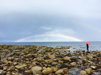 Person standing by sea with rainbow against sky during rain