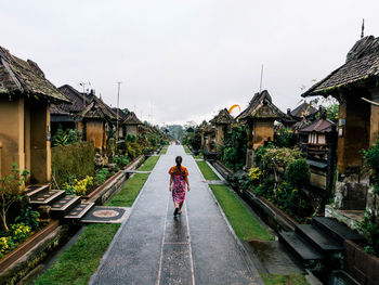 Rear view of woman walking on footpath amidst buildings against cloudy sky