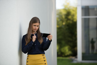 Thoughtful woman using mobile phone while standing by concrete wall