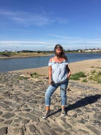 Woman with tousled hair standing at riverbank against blue sky