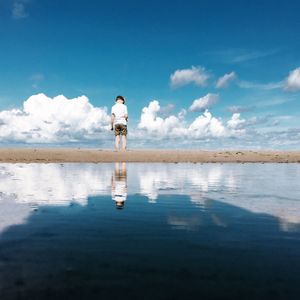 Rear view of boy standing at lakeshore against cloudy sky