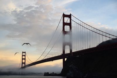 Low angle view of suspension bridge against cloudy sky