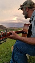 Side view of man playing guitar while sitting on land during sunset