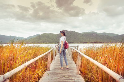 Woman standing on footbridge over lake against sky