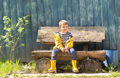 A little boy in a yellow jacket and rubber boots sits on a bench near a wooden fence. 