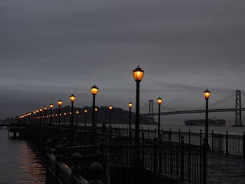 Illuminated street lights by sea against sky during sunset