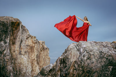 Low angle view of woman in red dress standing on rocky mountain against sky