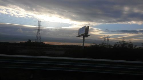 Electricity pylons on road against cloudy sky